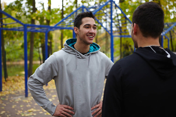 Lächelnder junger Sportler mit den Händen auf den Hüften im Gespräch mit seinem Freund während einer kurzen Trainingspause, Taillenbild - Foto, Bild