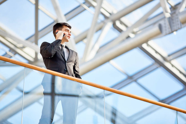 Low angle portrait of powerful young businessman talking by smartphone standing on glass balcony of modern office building - Foto, Imagem