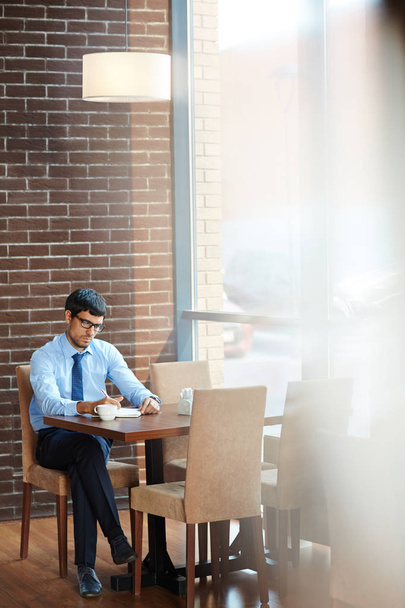 Γενειοφόρος υπαλληλικός εργαζόμενος σε γυαλιά που εργάζεται στο έργο του ενώ κάθεται στο ευρύχωρο καφέ με πανοραμικά παράθυρα - Φωτογραφία, εικόνα