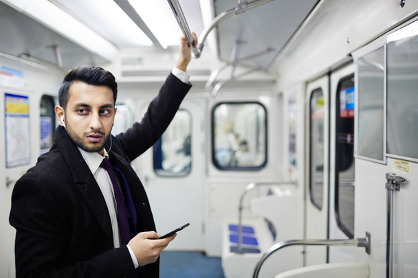 Portrait of handsome Middle-Eastern businessman standing in subway train holding onto hand rail and listening to music from smartphone - Photo, Image