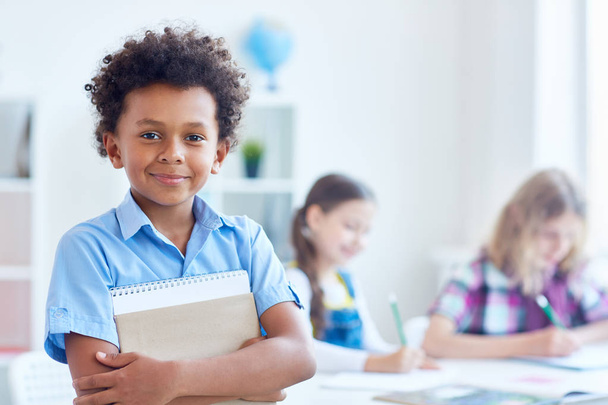 Cute schoolboy with copybook looking at camera with his classmates on background - Fotó, kép
