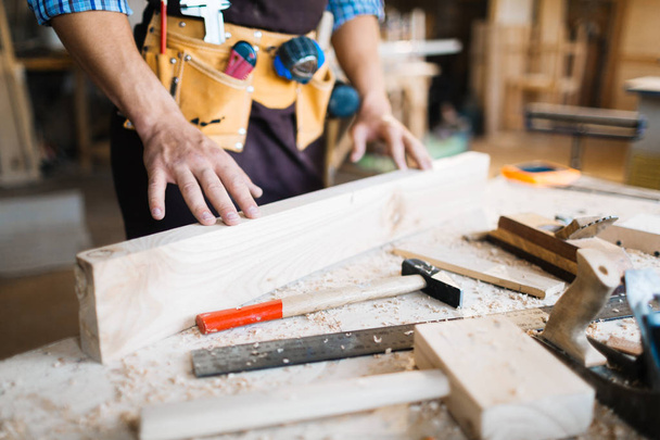 Nahaufnahme männlicher Hände, die die Oberfläche von Holzbrettern, Hämmern, Hobeln und Sägen untersuchen, die auf dem Tisch liegen - Foto, Bild