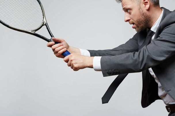 Vue latérale de l'homme d'âge moyen en costume d'affaires tenant la raquette de tennis prête à frapper la balle sur fond gris
 - Photo, image