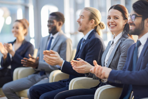 Multi-ethnic group of smiling business people sitting in row in modern glass hall and clapping, focus on beautiful brunette businesswoman looking at camera - Photo, Image