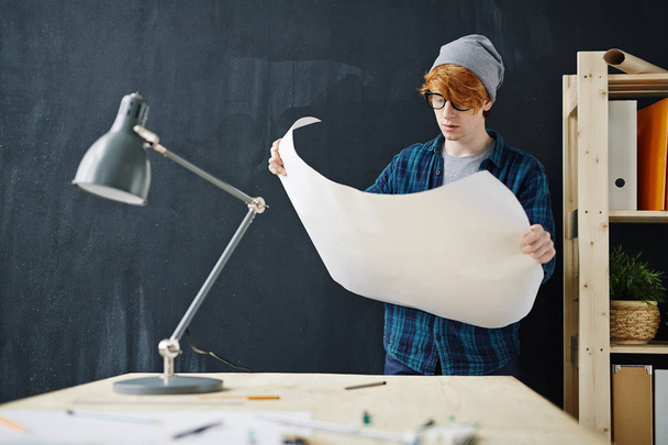 Portrait de jeune ingénieur aux cheveux rouges créatif portant un bonnet et des lunettes debout au bureau tout en travaillant avec des plans sur fond de tableau noir au bureau
 - Photo, image