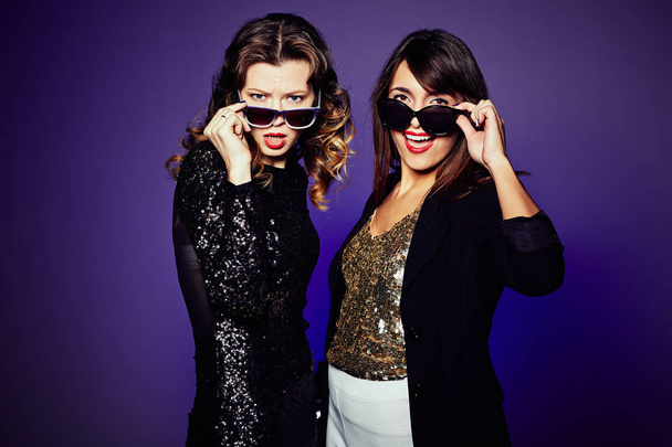 Две привлекательные модели позируют для фотографии в стильных солнцезащитных очках, стоя на тёмном фоне, студийный снимок
 - Фото, изображение