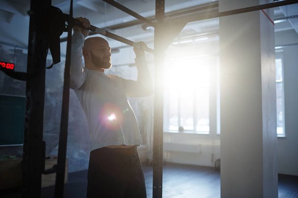 Portrait of strong bearded man performing pull ups on bar during workout in gym lit by sunlight - Fotoğraf, Görsel