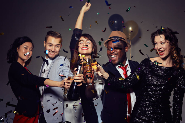 Met Nieuwjaar feest in discotheek: multi-etnische groep vrienden dansen en poseren voor fotografie met champagne fluiten in handen, kleurrijke confetti vallen op achtergrond - Foto, afbeelding