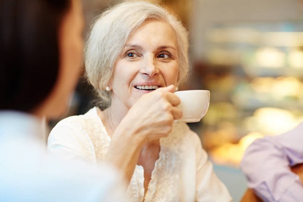 Зрелая женщина пьет чай из белого фарфора чашку, разговаривая с компаньоном
 - Фото, изображение