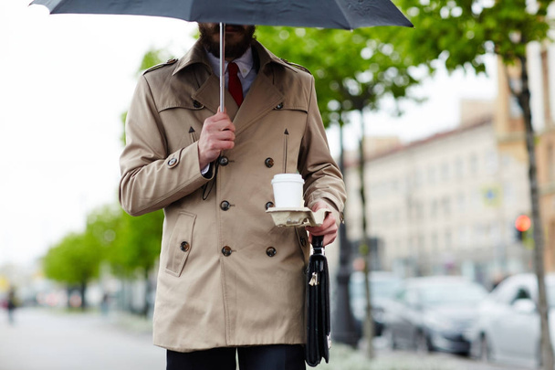 Businessman under umbrella carrying drink and briefcase - Photo, Image