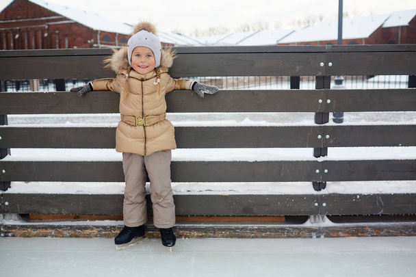 Criança feliz em winterwear em pé por cerca na pista de patinação
 - Foto, Imagem