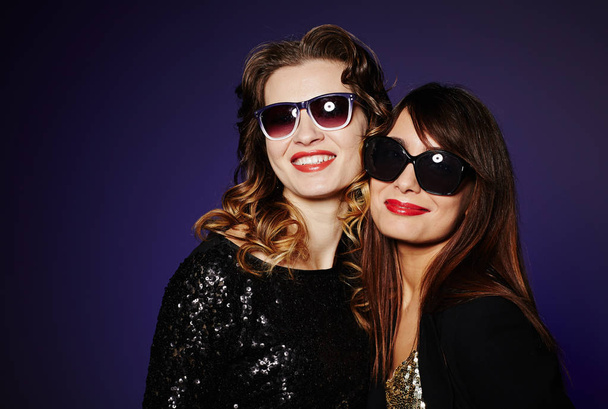 Μέση-κατακόρυφα δύο κομψά γυναικών σε γυαλιά ηλίου βλέπουν φωτογραφική μηχανή με ευρεία χαμόγελα ενώ στέκεται ενάντια σε σκούρο φόντο, studio που γυρίστηκε - Φωτογραφία, εικόνα