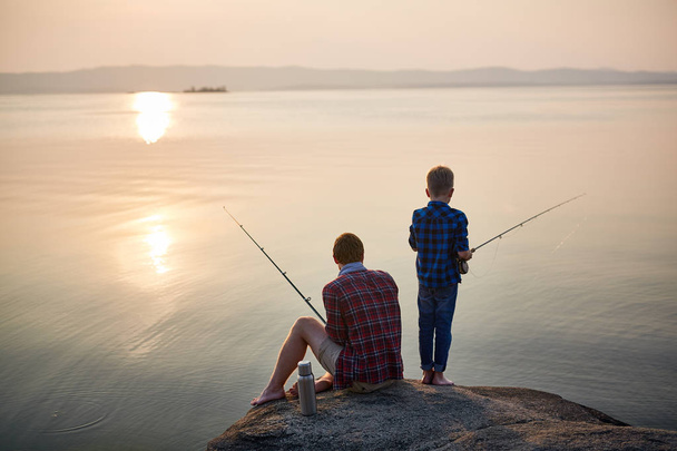 Back view portrait of adult man and teenage boy sitting together on rocks fishing with rods in calm waters with landscape of setting sun, both wearing checkered shirts - Photo, Image