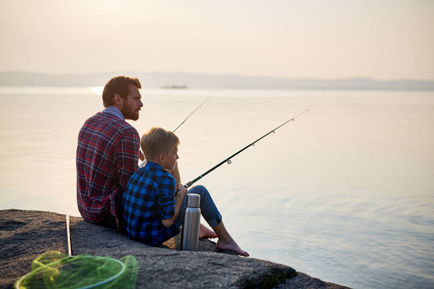 Portrait de l'homme adulte et adolescent assis ensemble sur des rochers pêchant avec des cannes dans les eaux calmes du lac bleu au crépuscule, tous deux portant des chemises à carreaux
 - Photo, image