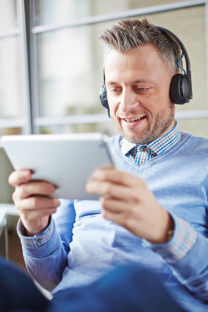 Curious middle-aged businessman in headphones using Internet on tablet to watch online seminar - Photo, image