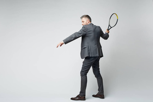 Retrato de comprimento total de empresário de meia-idade em terno bater tiro com raquete de tênis contra fundo cinza
 - Foto, Imagem