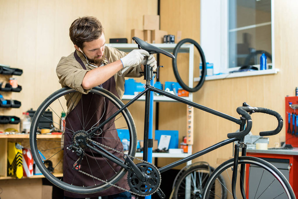 Bearded young mechanic wearing apron listening to music in headphones while adjusting bicycle saddle at modern workshop - Foto, Imagem