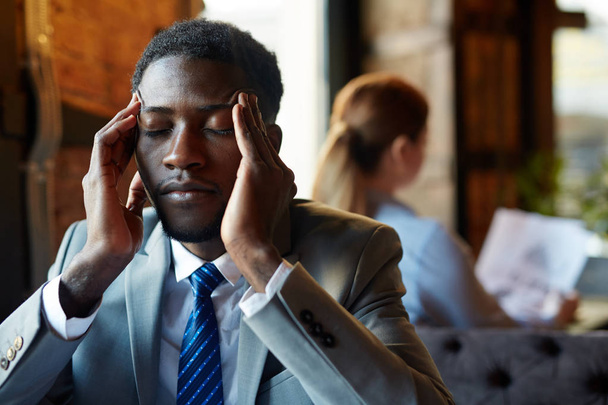 Retrato de un hombre afroamericano guapo con traje de negocios frotándose las sienes y cerrando los ojos sufriendo de dolor de cabeza y agotamiento en la mesa en el restaurante moderno
  - Foto, Imagen