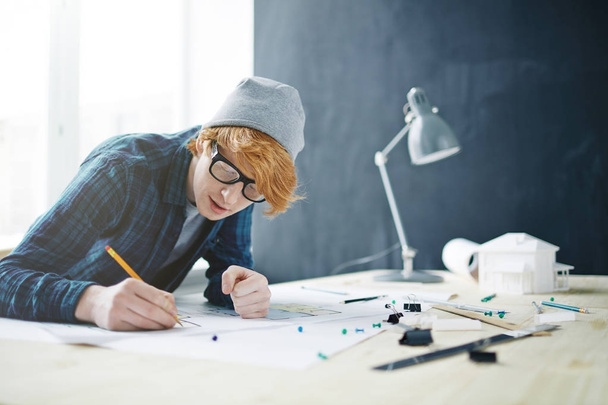 Portrait of young red haired designer wearing glasses and beanie hat working in office: leaning on desk with lamp and house model, drawing floor plan - Photo, image