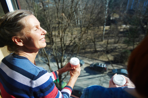 Ευτυχισμένη γυναίκα με ζεστό ποτό, απολαμβάνοντας το πρωινό ήλιο στο ανοιχτό παράθυρο - Φωτογραφία, εικόνα