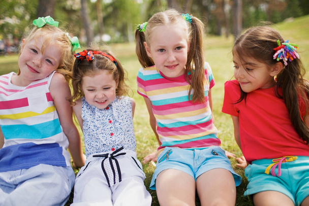 Yaz aylarında neşeli küçük kızlar yakından görmek Park yeşil çimenlerin üzerinde oturan ve kameraya bakarak elbise - Fotoğraf, Görsel