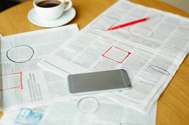 Krant met gemarkeerde vacature-advertenties, smartphone, kopje koffie en pen op tafel - Foto, afbeelding