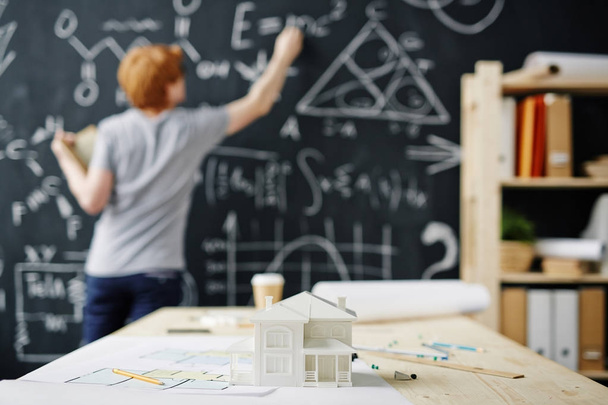 家モデルと背景に黒板に数式を書く若い建築家の机の上のフロア プラン - 写真・画像