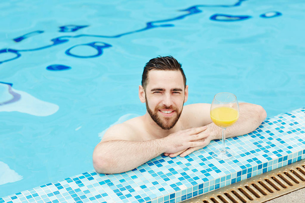 Happy topless man with glass of drink looking out of swimming-pool by hotel of spa resort - Photo, Image