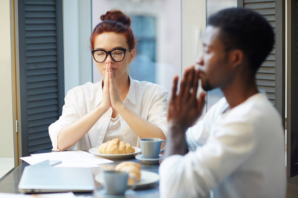 Aantrekkelijke jonge vrouw en haar knappe Afro-Amerikaanse vriend verzameld rond tafel en bidden voordat u ontbijt, huisgemaakte croissants en kruidenthee te wachten voor ze - Foto, afbeelding