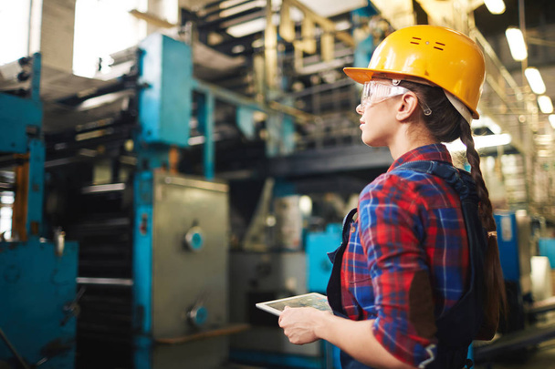 Ingeniero trabajador sosteniendo la tableta digital en las manos mientras trabaja en la planta de fabricación, vista de perfil
 - Foto, Imagen