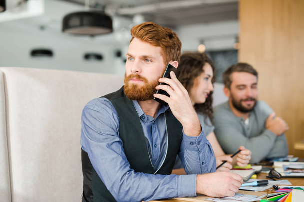 Young bearded business leader speaking on mobile phone with his team behind - Fotoğraf, Görsel