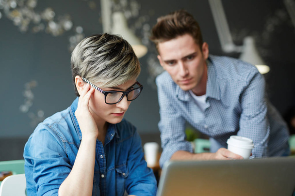 Portrait of young businesswoman wearing creative short haircut and glasses looking confused while working with laptop in office, colleague looking at screen beside her - Fotoğraf, Görsel