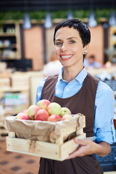 schwarzhaarige Ladenbesitzerin hält kleine Holzkiste mit reifen Äpfeln in den Händen, während sie für Fotos posiert, verschwommener Hintergrund - Foto, Bild