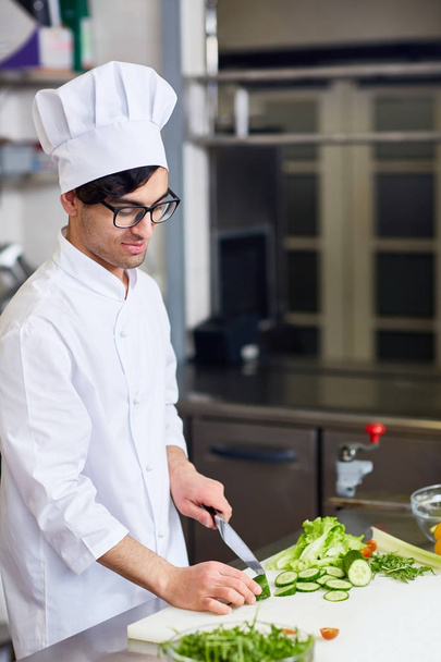 Young man in chef uniform cutting fresh cucumber for salad - Photo, Image