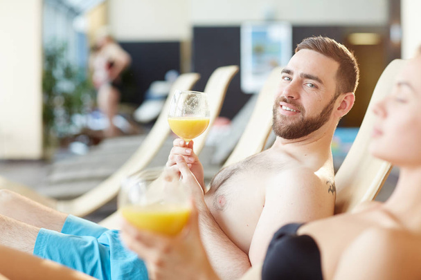 Przystojny mężczyzna z szklanka soku, patrząc na kamery podczas relaksu w spa resort ze swoją dziewczyną w pobliżu przez - Zdjęcie, obraz