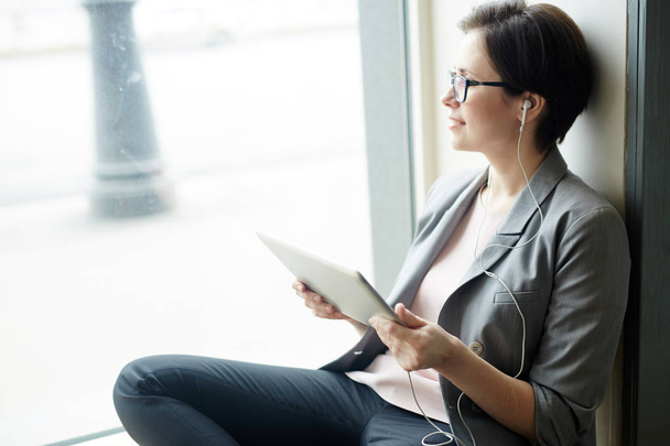 Νεαρή γυναίκα με το touchpad, ακούγοντας μουσική και αναζητούν μέσα από το παράθυρο - Φωτογραφία, εικόνα