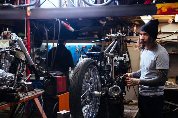 Side view portrait of bearded tattooed man working in garage tuning  motorcycle and repairing broken parts - Photo, Image