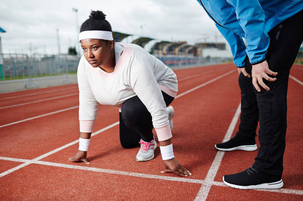 Jovem mulher desportiva de grandes dimensões em pé pela linha de início pronto para correr com seu treinador por perto
 - Foto, Imagem