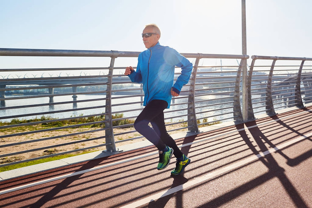 Здоровий літній чоловік у спортивному одязі бігає по спортивній трасі, готуючись до марафону, мальовничий вид на тлі, безхмарне блакитне небо
 - Фото, зображення