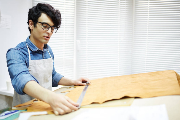 Young artisan taking measures of piece of fabric by workplace before cutting it - Φωτογραφία, εικόνα