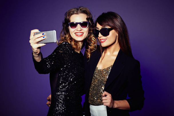 Attractive it girls in trendy sunglasses taking selfie on smartphone before starting to celebrate New Year, waist-up portrait - Photo, Image