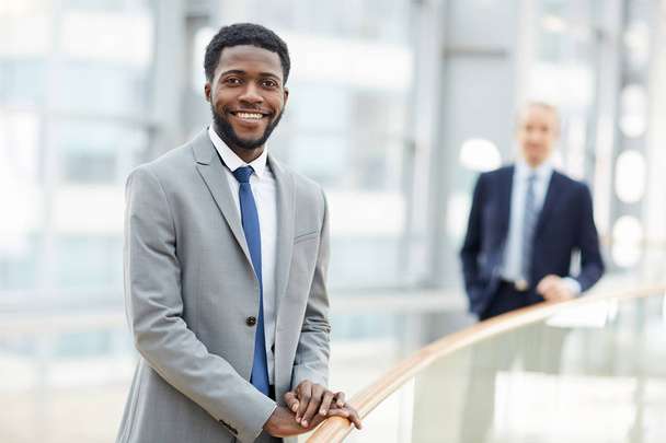 Retrato de um empresário afro-americano sorridente olhando para a câmera apoiada em trilhos de vidro no moderno prédio de escritórios, segundo homem em segundo plano
 - Foto, Imagem