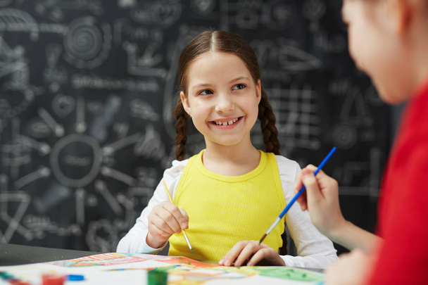 Portret ładny uśmiechający się mały dziewczyna malarstwo obrazów z przyjacielem podczas lekcji sztuki w szkole siedząc przed tablica - Zdjęcie, obraz