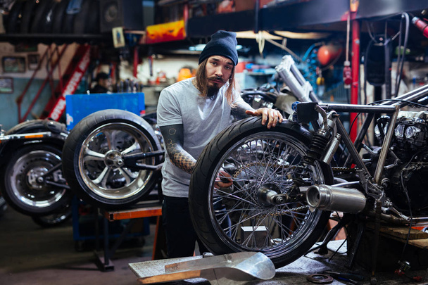 Portrét vousatého Tetovaný muž pracuje v garáži, tuning motocyklu a při pohledu na fotoaparát - Fotografie, Obrázek