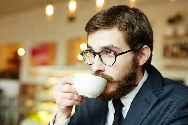 Fiatal férfi szemüvegek, fehér porcelán csésze teát vagy kávét iszik és fejrevalók - Fotó, kép