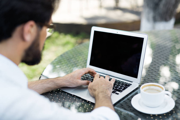 Over shoulder view of bearded businessman sitting in outdoor cafe and working on his modern laptop, empty cup of coffee standing on table - Photo, Image
