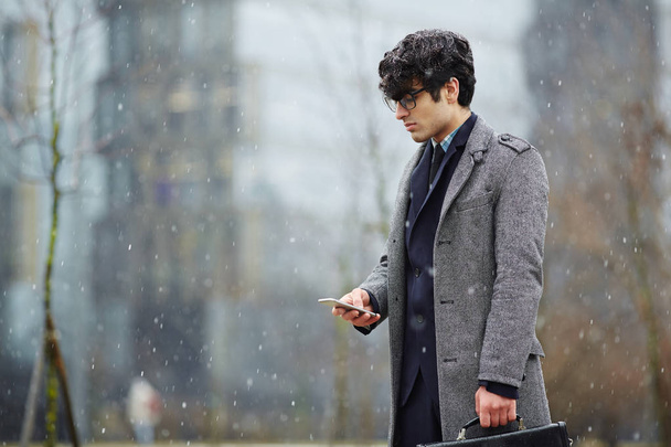 Портрет молодого ближневосточного бизнесмена, стоящего на улице в снегопаде и использующего смартфон, смотрящего на экран
 - Фото, изображение