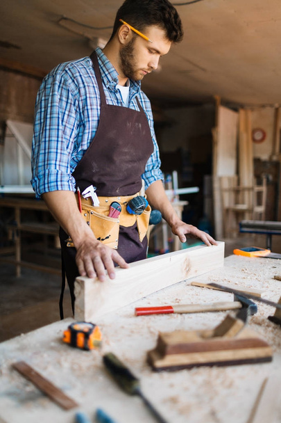 Vousatý carpenter s tužkou za uchem Kontrola povrchu dřevěné desky, stůl s hoblinami a ruční nástroje na popředí - Fotografie, Obrázek