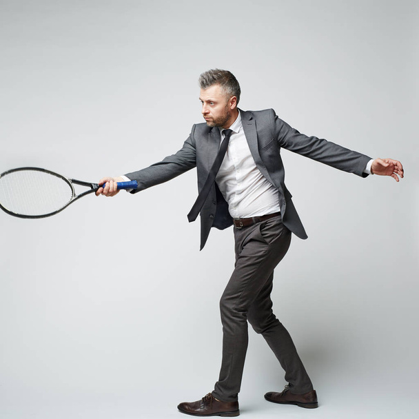 Middle aged grey haired businessman in suit imitating tennis game against grey background - Photo, image