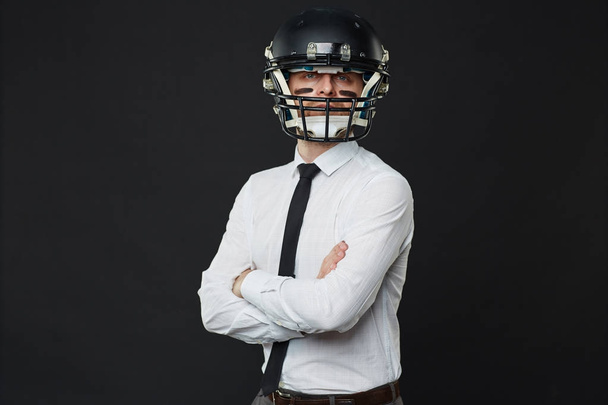 Waist up portrait of confident businessman wearing American football helmet standing cross-armed against black background and looking at camera - Photo, Image